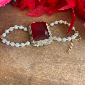 Puede incluir: Un brazalete de oro con una gran piedra preciosa roja y detalles de perlas blancas.