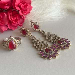 May include: A set of silver earrings and a ring with red and purple gemstones. The earrings feature a teardrop-shaped red stone surrounded by smaller purple stones and a row of clear stones. The ring has a similar teardrop-shaped red stone surrounded by clear stones.