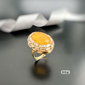 May include: A gold ring featuring a large, oval-shaped orange gemstone, surrounded by a band of smaller, rectangular clear stones. The ring has a delicate gold band and is set against a dark background. The ring weighs 8.9 grams.