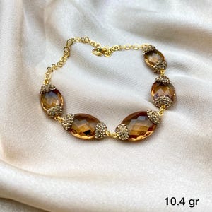 May include: A gold-toned bracelet featuring oval-shaped, amber-colored gemstones. Each gemstone is accented with small, sparkling embellishments. The bracelet is displayed on a white, silky fabric. The text "10.4 gr" is visible in the image.