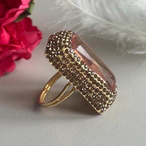 May include: A gold ring with a large, rectangular, faceted, pink gemstone. The gemstone is surrounded by a border of small, brown rhinestones.
