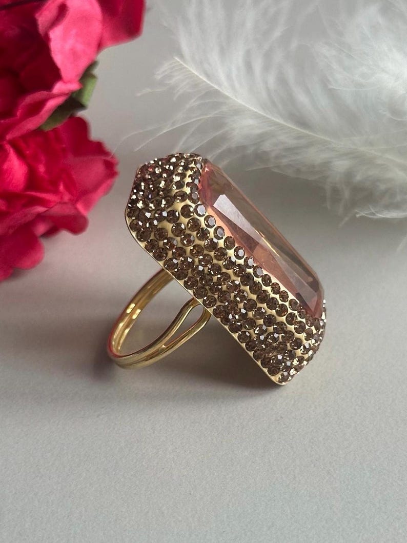May include: A gold ring with a large, rectangular, faceted, pink gemstone. The gemstone is surrounded by a border of small, brown rhinestones.