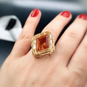 Peut inclure: Une bague dorée ornée d'une grande pierre ambre rectangulaire au centre, entourée de petites pierres rectangulaires transparentes. La bague est portée sur une main avec du vernis à ongles rouge.