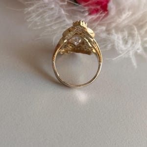 May include: A gold ring with a large clear stone set in a decorative design. The ring is on a white background.