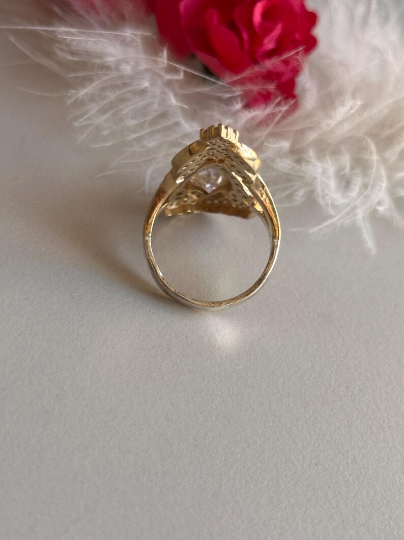 May include: A gold ring with a large clear stone set in a decorative design. The ring is on a white background.