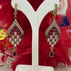 May include: A pair of dangle earrings with a teardrop-shaped design. The earrings are made of gold-tone metal and feature clear and red cubic zirconia stones.