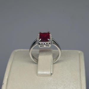 May include: A silver ring with a square, deep red gemstone, encircled by a halo of smaller, clear stones. The ring's band is smooth and polished, creating a classic design. The ring is displayed on a neutral-toned surface.