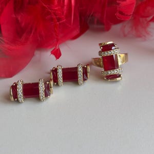 May include: A set of gold-tone jewelry with red gemstone accents. The set includes a ring and two earrings. The ring has a large, rectangular red gemstone surrounded by smaller clear gemstones. The earrings are similar in design, with smaller rectangular red gemstones.