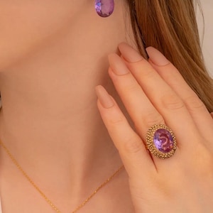 Puede incluir: Un conjunto de joyas doradas con un anillo, pendientes y un collar. El anillo está en un dedo y los pendientes en una oreja. Cada pieza tiene una gran piedra preciosa ovalada púrpura con un engaste dorado texturizado. El collar tiene un colgante a juego.