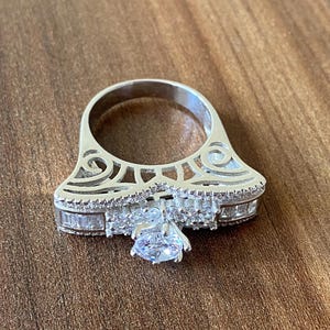 May include: A silver ring with a large, clear, round-cut center stone surrounded by smaller, baguette-cut stones. The ring has a delicate, intricate design with a filigree pattern.
