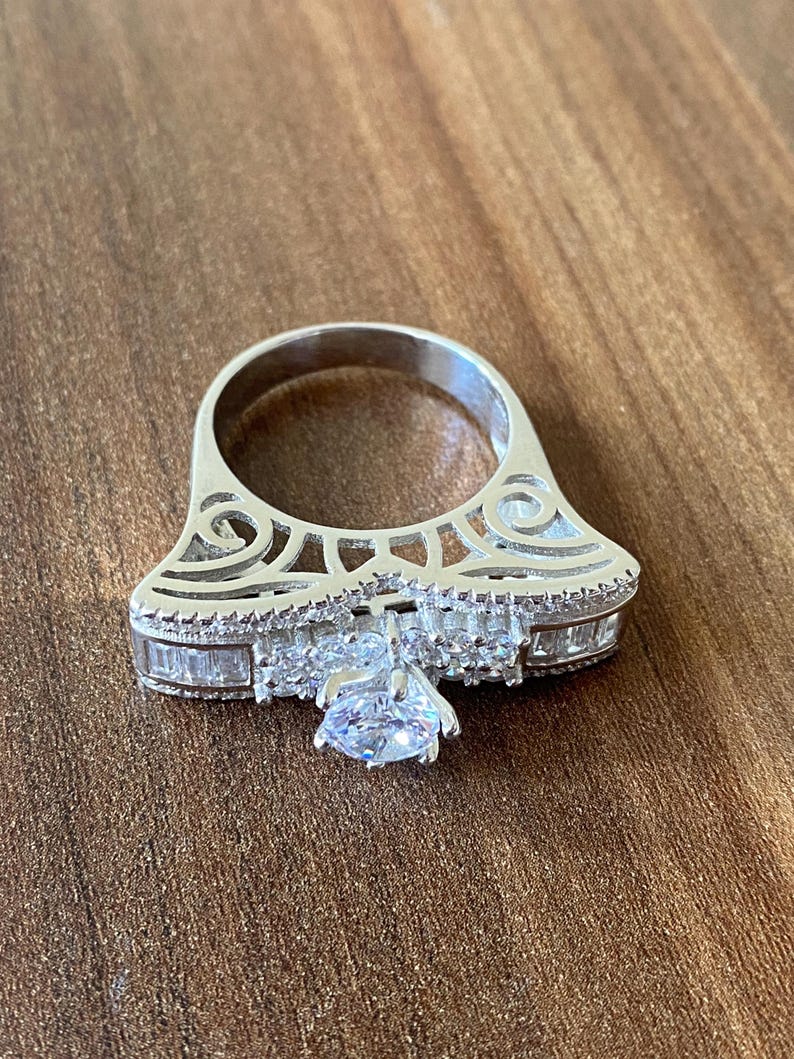 May include: A silver ring with a large, clear, round-cut center stone surrounded by smaller, baguette-cut stones. The ring has a delicate, intricate design with a filigree pattern.
