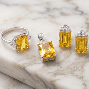 May include: A jewelry set consisting of a silver-toned ring, a pendant, and a pair of earrings. Each piece features a large, rectangular, yellow gemstone flanked by smaller, clear, triangular stones set in a metallic frame.