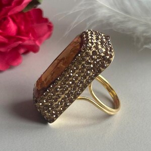 May include: A gold ring with a large, rectangular, faceted stone set in the center. The stone is covered in small, clear rhinestones.