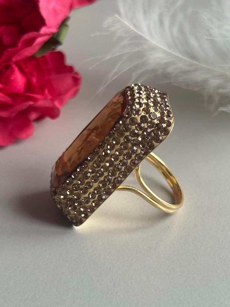 May include: A gold ring with a large, rectangular, faceted stone set in the center. The stone is covered in small, clear rhinestones.