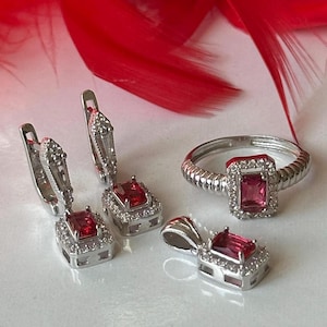 Peut inclure: Un ensemble de bijoux en argent avec des pierres précieuses rouges. L'ensemble comprend une bague, un pendentif et une paire de boucles d'oreilles. Chaque pièce présente une pierre précieuse rouge de forme carrée entourée de petites pierres précieuses claires.