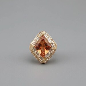 May include: A square-cut, orange gemstone ring with a halo of rectangular, clear stones. The ring's setting is gold-colored, and the design is set against a gray background. The ring is a statement piece.