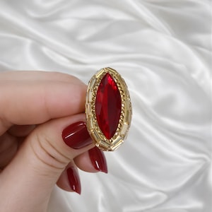 Può includere: Un anello dorato con una grande gemma rosso scuro di forma ovale. La gemma è incastonata in una montatura dorata strutturata. L'anello è presentato su uno sfondo bianco e serico.