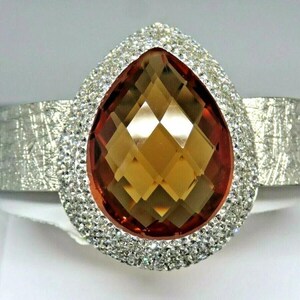 May include: A silver bracelet featuring a large, teardrop-shaped, faceted, orange-brown gemstone set within a halo of clear, sparkling stones.