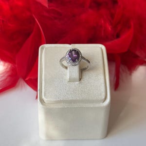 May include: A silver ring with a large oval, purple gemstone at the center, surrounded by a halo of smaller, clear stones. The ring is displayed on a white jewelry stand, with a backdrop of red feathers.