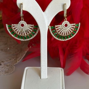 Könnte beinhalten: Goldene Ohrringe mit einem fächerförmigen Design, das mit klaren und grünen Zirkonia-Steinen besetzt ist.