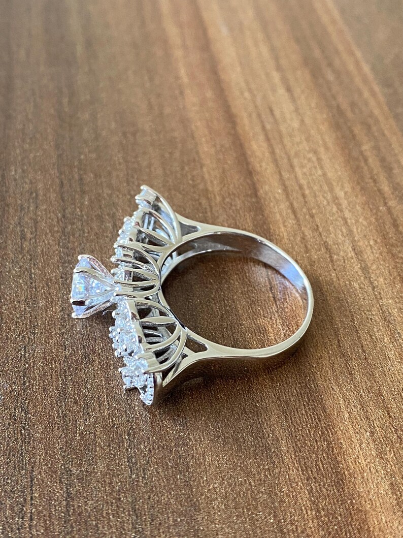 May include: A silver ring with a large, clear, round gemstone surrounded by smaller, clear gemstones. The ring has a delicate, intricate design.