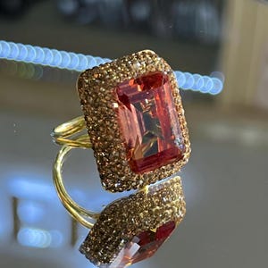 May include: A gold-toned ring featuring a large, rectangular, reddish-orange gemstone. The gemstone is surrounded by a border of smaller, sparkling, golden-brown stones. The ring's band is a simple, elegant design.