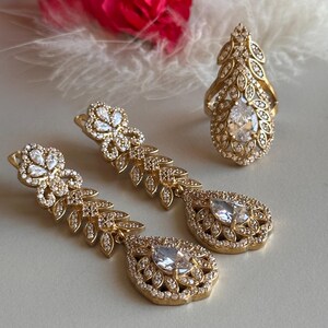 May include: A set of gold-tone jewelry featuring a ring and a pair of dangle earrings. The pieces are adorned with clear cubic zirconia stones and intricate leaf designs.