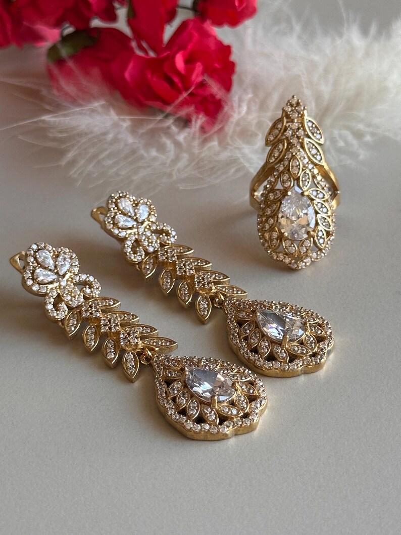 May include: A set of gold-tone jewelry featuring a ring and a pair of dangle earrings. The pieces are adorned with clear cubic zirconia stones and intricate leaf designs.
