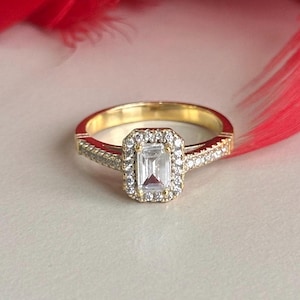May include: A gold ring with a large, clear, rectangular-shaped gemstone surrounded by smaller diamonds. The ring is set on a white background.