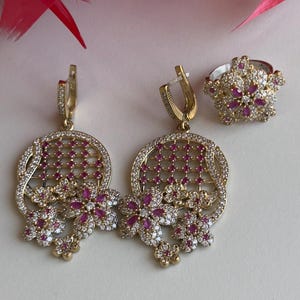 Può includere: Un paio di orecchini d'oro con pietre rosa e bianche. Gli orecchini presentano un design circolare con un motivo a traliccio e fiori pendenti. È incluso anche un anello abbinato con un design simile.