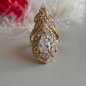May include: A gold ring with a large pear-shaped clear stone surrounded by smaller clear stones. The ring has a leaf-like design.