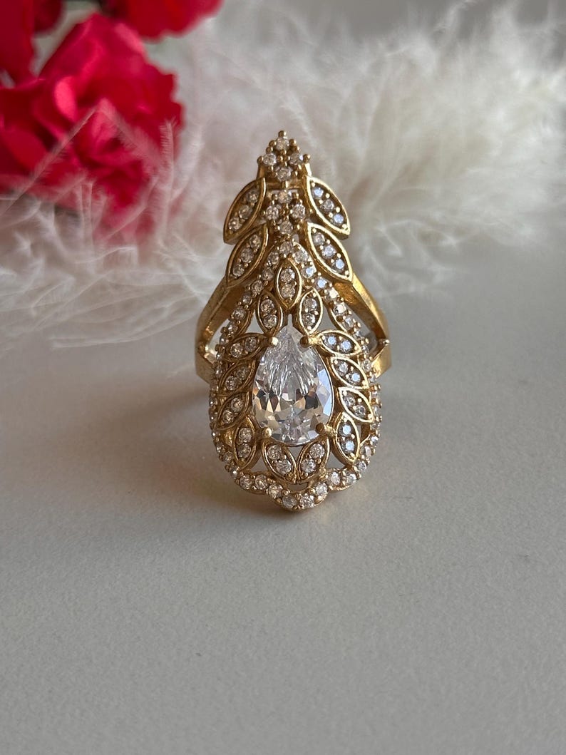 May include: A gold ring with a large pear-shaped clear stone surrounded by smaller clear stones. The ring has a leaf-like design.