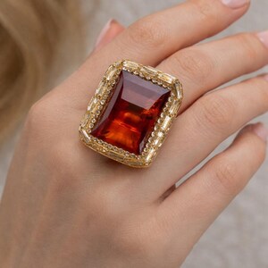 May include: A large, rectangular cocktail ring with a gold setting. The ring features a large, faceted, reddish-brown gemstone surrounded by smaller, clear stones. The ring is on a person's hand.