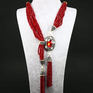 May include: A long red beaded necklace with a large, oval-shaped pendant featuring a red gemstone surrounded by smaller, clear stones. The necklace has two tassels at the bottom.