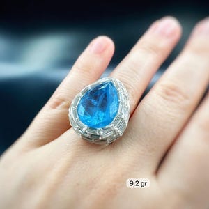 May include: A large, eye-catching ring featuring a pear-shaped, light blue gemstone. The gemstone is surrounded by a halo of smaller, rectangular, clear stones. The ring is set on a silver band. The weight of the ring is 9.2 grams.