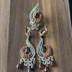 Puede incluir: Un conjunto de pendientes y un anillo con una piedra roja y piedras de circonita cúbica transparentes. Las joyas son de color dorado y tienen un diseño delicado e intrincado.