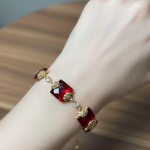 Pode incluir: Uma pulseira dourada com tr&ecirc;s pedras preciosas quadradas vermelhas escuras facetadas. A pulseira &eacute; conectada por elos dourados menores com pedras de acento transparentes. A pulseira &eacute; usada em um pulso.