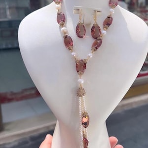 Puede incluir: Un conjunto de joyas que incluye un collar, una pulsera y pendientes. El conjunto incluye grandes piedras preciosas rosas de forma ovalada, pequeñas perlas blancas y detalles dorados. El collar tiene un diseño largo y colgante.