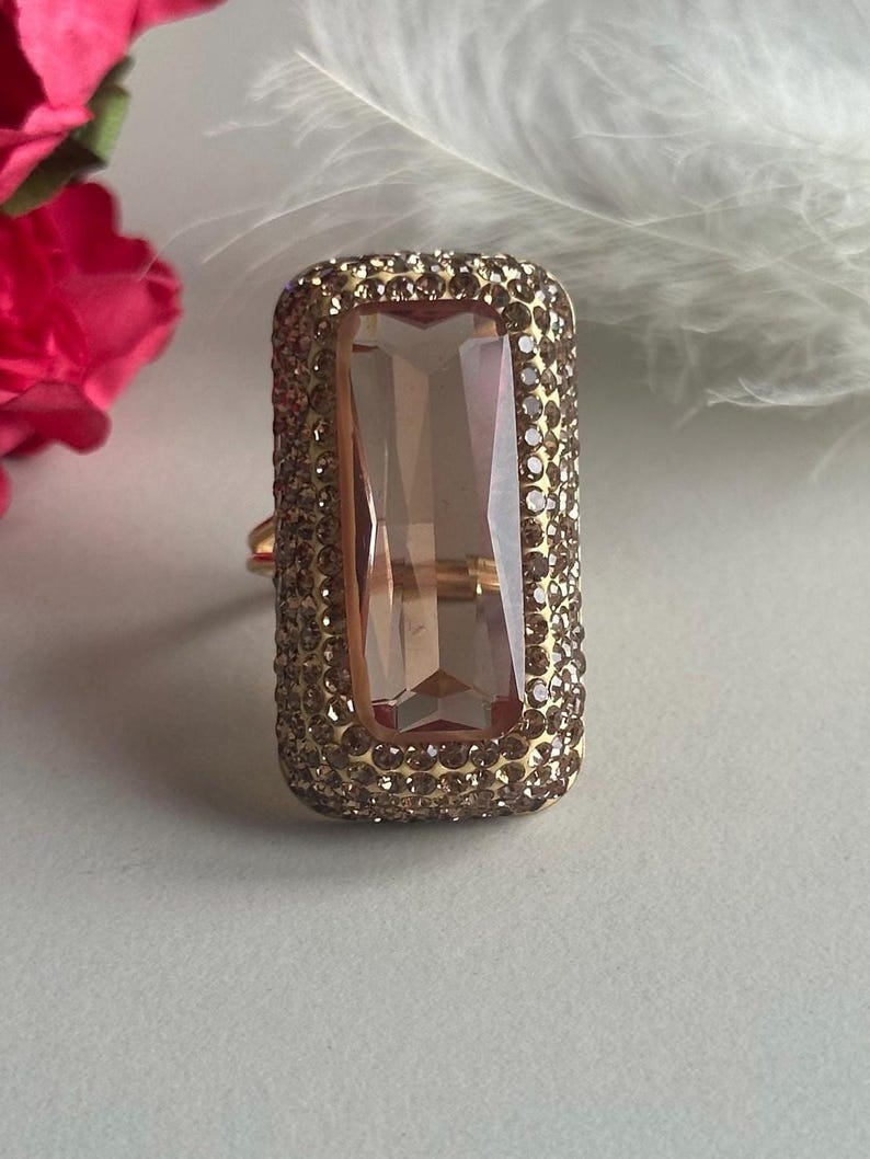 May include: A gold ring with a large, rectangular, clear pink gemstone surrounded by smaller clear stones.