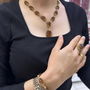 Puede incluir: Un conjunto de joyas doradas que incluye un collar, una pulsera y un anillo con piedras preciosas marrones. El collar tiene un colgante en forma de lágrima. La pulsera y el collar tienen un diseño similar. También se ve un reloj dorado.