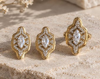 Könnte beinhalten: Ein Goldschmuckset bestehend aus zwei Ohrsteckern und einem Ring. Jedes Stück verfügt über einen zentralen, klaren Stein im Marquise-Schliff, umgeben von einem Kranz aus kleinen Pavé-Steinen in einer geschwungenen, länglichen Goldfassung.