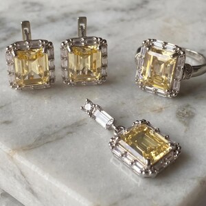 May include: A jewelry set consisting of a pair of earrings, a ring, and a pendant. Each piece features a large, rectangular, yellow center stone surrounded by a halo of small, clear, baguette-cut stones set in a silver-toned metal frame.