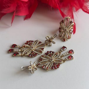 Puede incluir: Un conjunto de pendientes y un anillo de color dorado con piedras rojas y transparentes. Los pendientes son de estilo colgante con un diseño floral y el anillo es una banda ancha con un diseño floral.