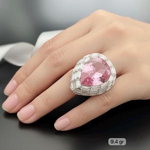 May include: A statement ring with a large, pink, pear-cut gemstone, encircled by a border of rectangular stones. The ring is set in silver or white metal. The ring weighs 9.4 grams.
