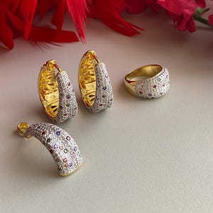 May include: A set of gold-toned jewelry featuring a ring, a pair of hoop earrings, and a pendant. All pieces are adorned with small, colorful gemstones and clear cubic zirconia stones.