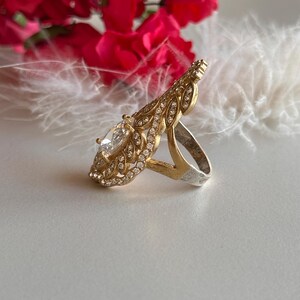 May include: A gold ring with a large clear stone surrounded by smaller stones. The ring has a delicate, leaf-like design.