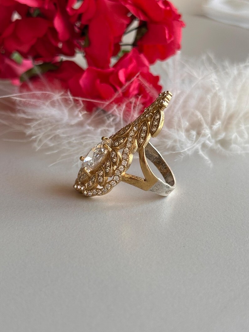 May include: A gold ring with a large clear stone surrounded by smaller stones. The ring has a delicate, leaf-like design.