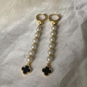 May include: A pair of gold-toned hoop earrings with a dangling design. Each earring features a strand of white faux pearls, gold accents, and a black clover charm with a gold border. The earrings are displayed on a white, textured surface.