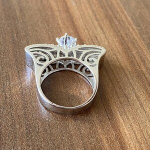 May include: A silver ring with a large, clear, round stone set in a filigree design. The ring is on a wooden surface.