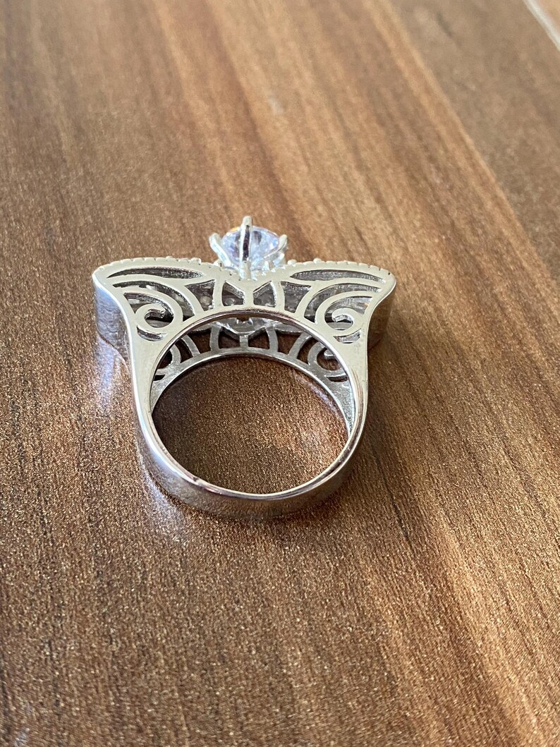 May include: A silver ring with a large, clear, round stone set in a filigree design. The ring is on a wooden surface.